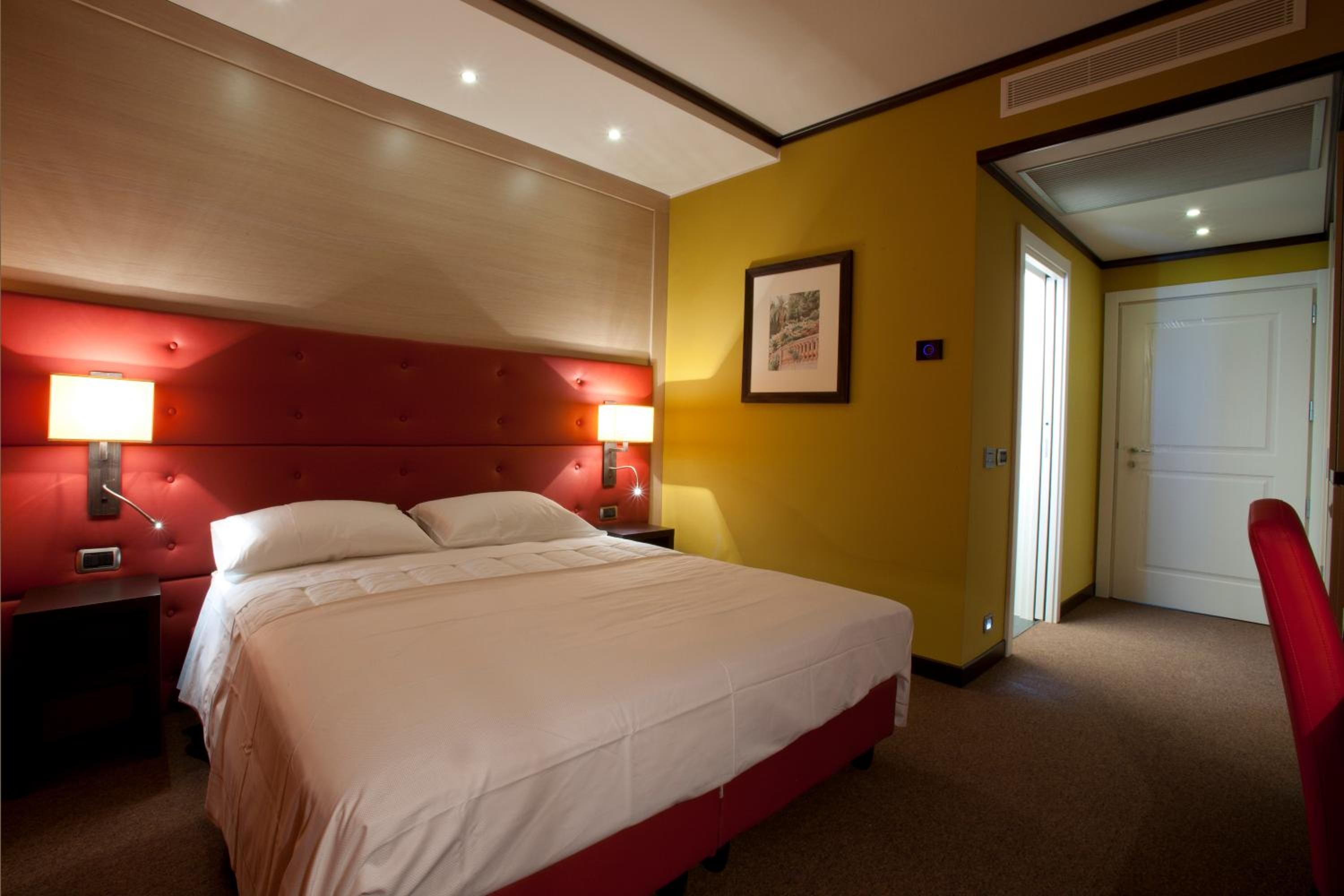 A hotel room with a red upholstered headboard, a large bed with white linens, two bedside lamps, and a framed picture on the wall.