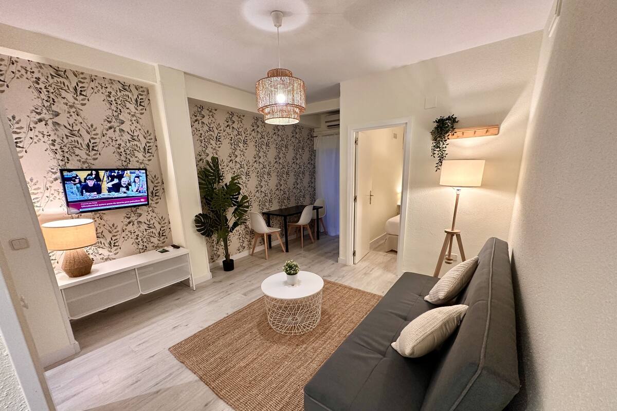 A modern living room with a sofa, coffee table, and TV. There is a dining area with chairs and a table in the background.