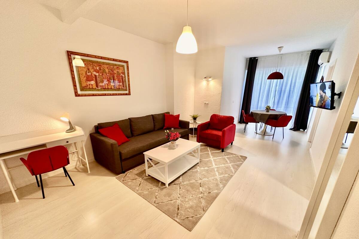 A modern living room with a sofa, red chairs, a coffee table, and a dining area with a table and chairs.