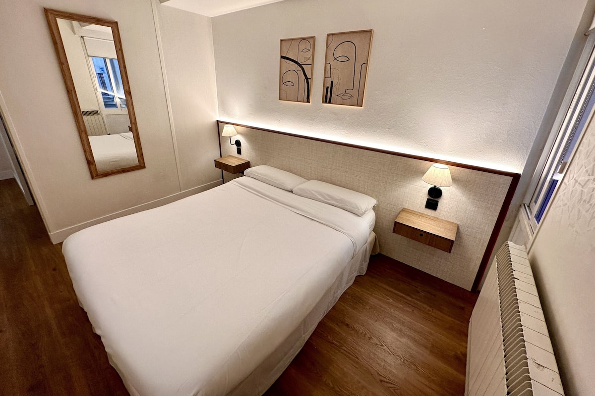 A hotel room with a large bed, a nightstand with a lamp, a mirror, and two wall-mounted artworks.