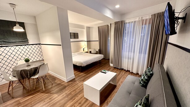 A modern hotel room with a bed, a sofa, a dining area with a table and chairs, a TV mounted on the wall, and a window with curtains. - Far Home Gran Vía - Hostel (Madrid)