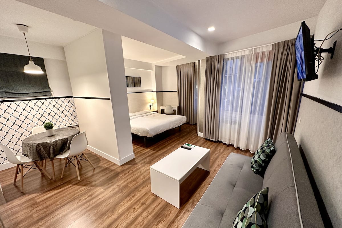 A modern hotel room with a bed, a sofa, a dining area with a table and chairs, a TV mounted on the wall, and a window with curtains.