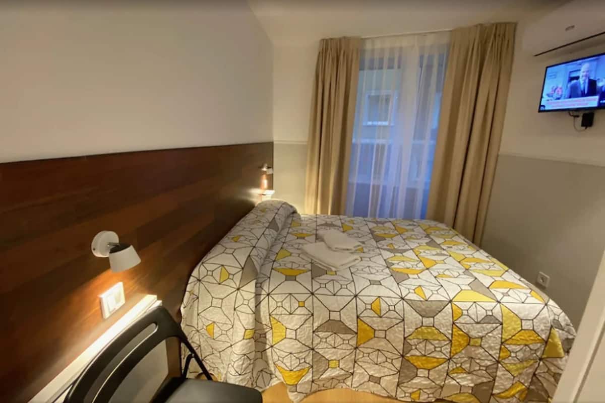 A hotel room with a wooden headboard, a bed with a geometric patterned bedspread, a television on the wall, and a window with curtains.