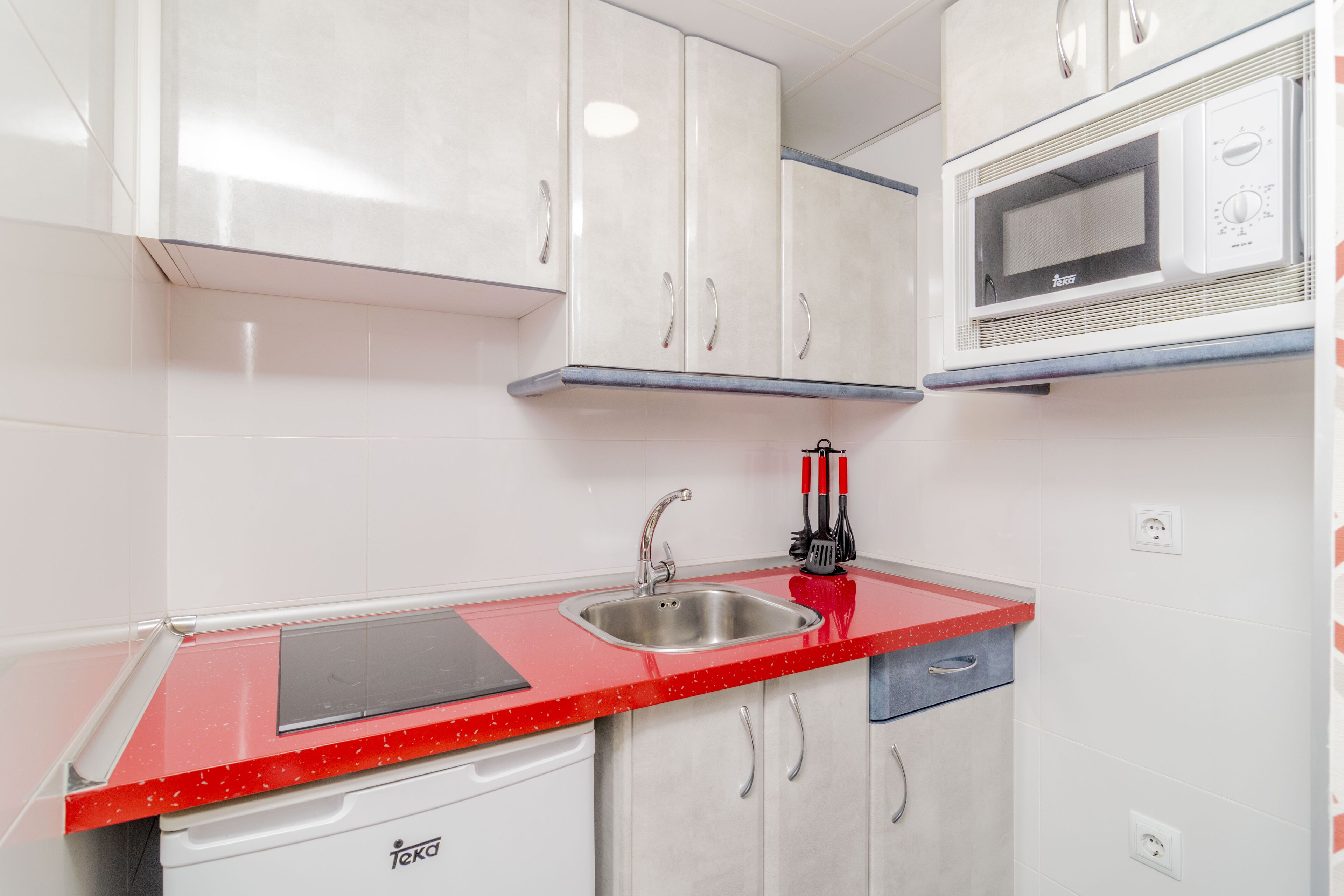 A compact kitchen with white cabinets, a red countertop, a sink, and a microwave.