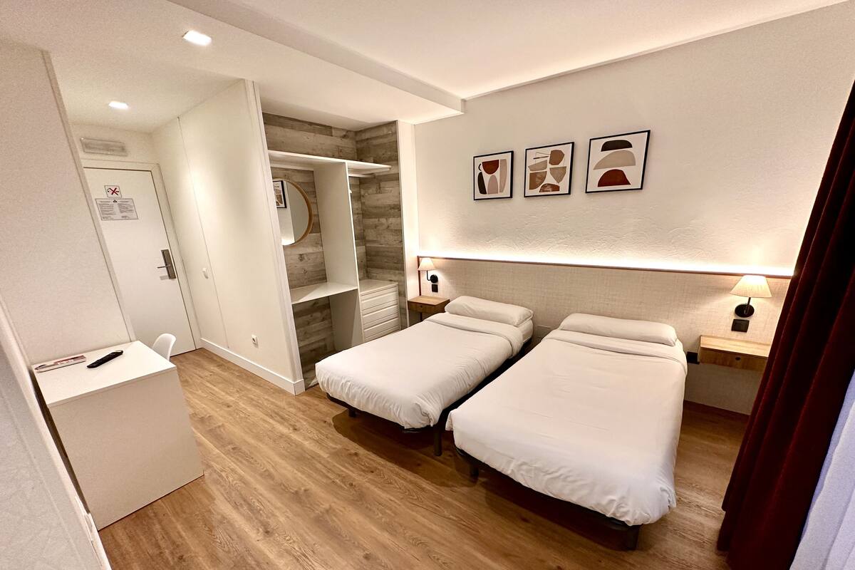 A hotel room with two single beds, a wooden floor, a wardrobe, and a small desk with a lamp.