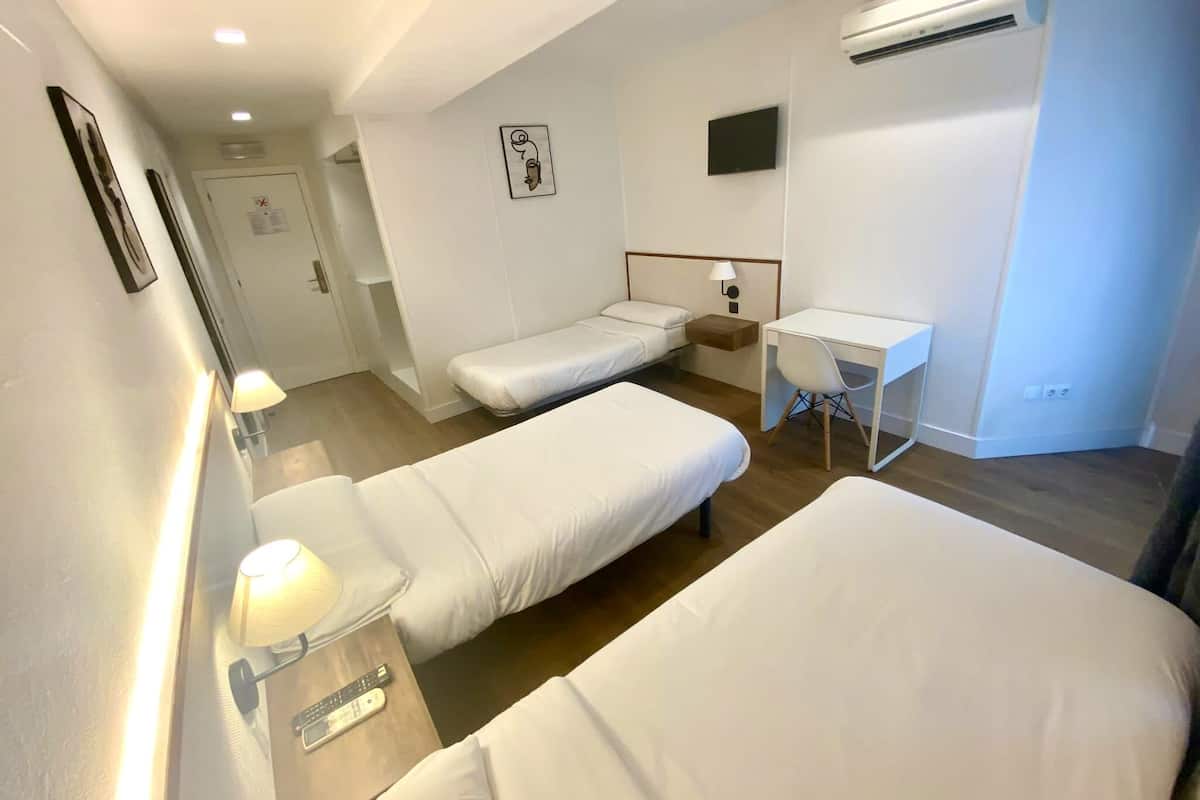 A hotel room with two single beds, a desk, a chair, a TV, and an air conditioning unit.