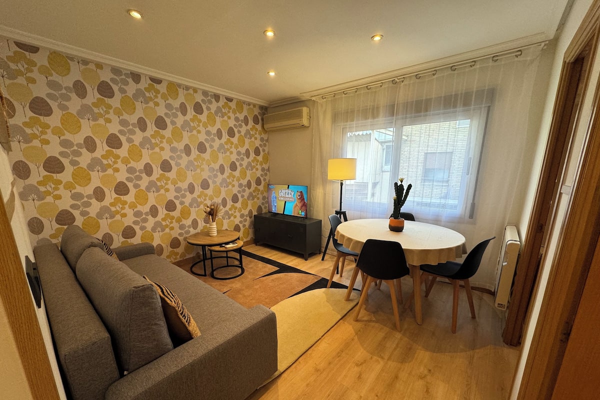 A living room with a sofa, a coffee table, and a dining table with chairs. There is a TV on a stand, a lamp, and a cactus on the table.