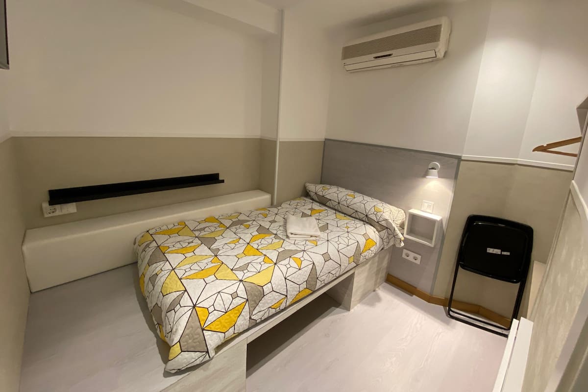 A small, single-bed room with a geometric-patterned bedspread, a bedside table with a lamp, a black trash bin, and a wall-mounted air conditioner.