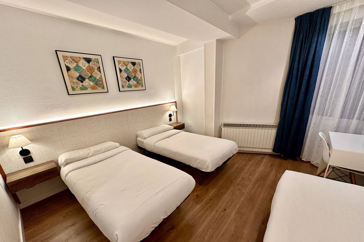 A hotel room with two single beds, a desk, a chair, a lamp, and two framed artworks on the wall.