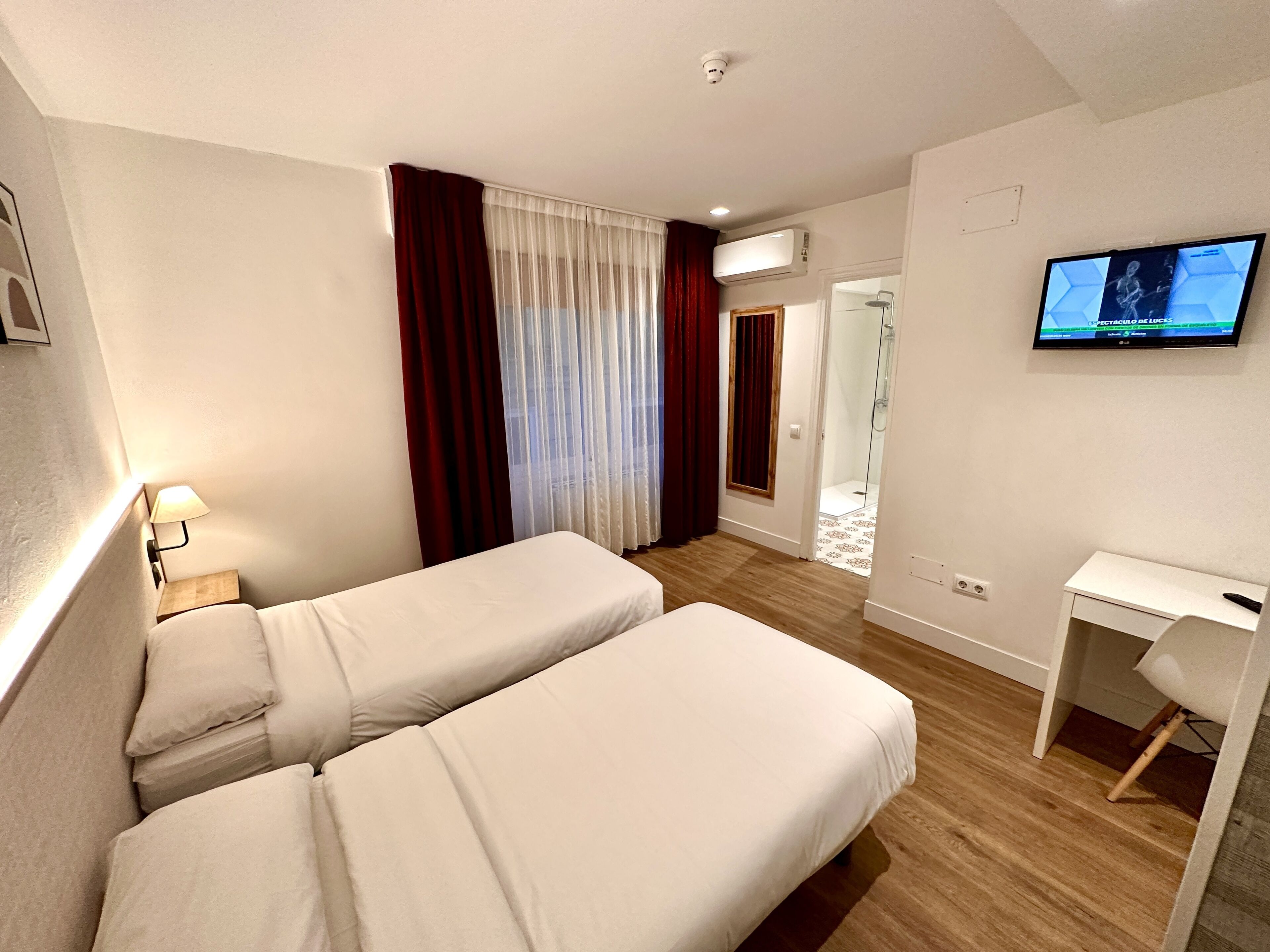 A hotel room with two beds, a desk, a TV, and a shower area.
