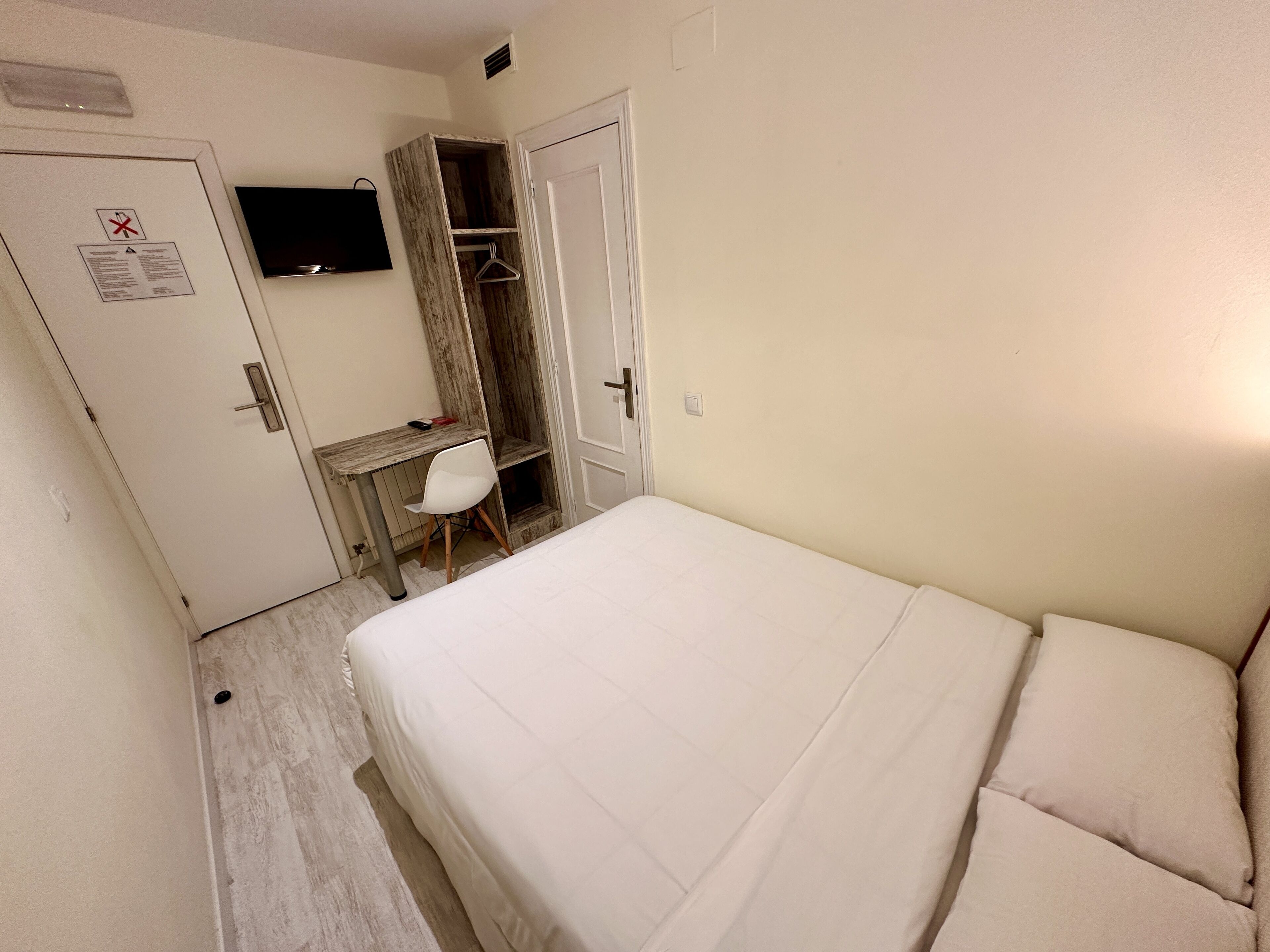 A small, clean room with a bed, a desk, and a chair. There is a door with a sign on it and a television mounted on the wall.