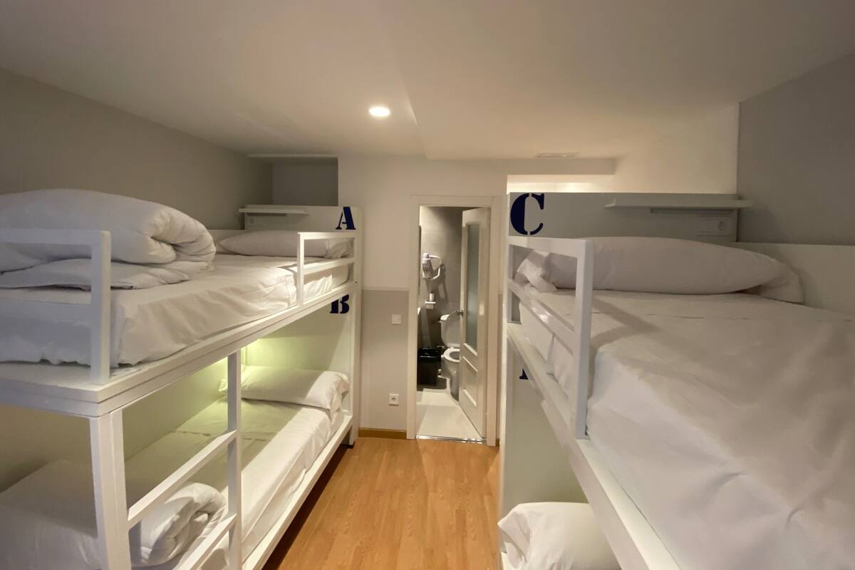 A dormitory-style room with bunk beds, a wooden floor, and a small bathroom visible through an open door.