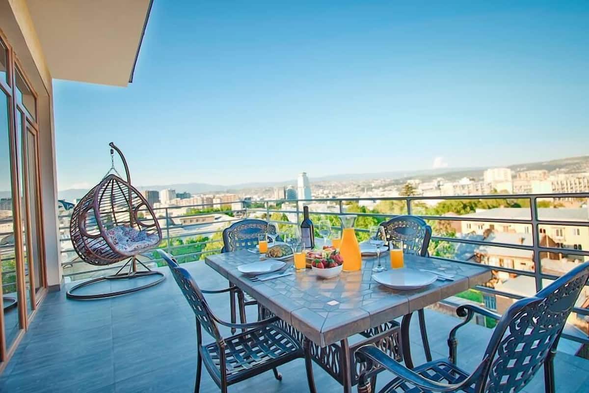 Un balcón con una mesa lista para dos, sillas, una hamaca colgante y una botella de zumo de naranja.