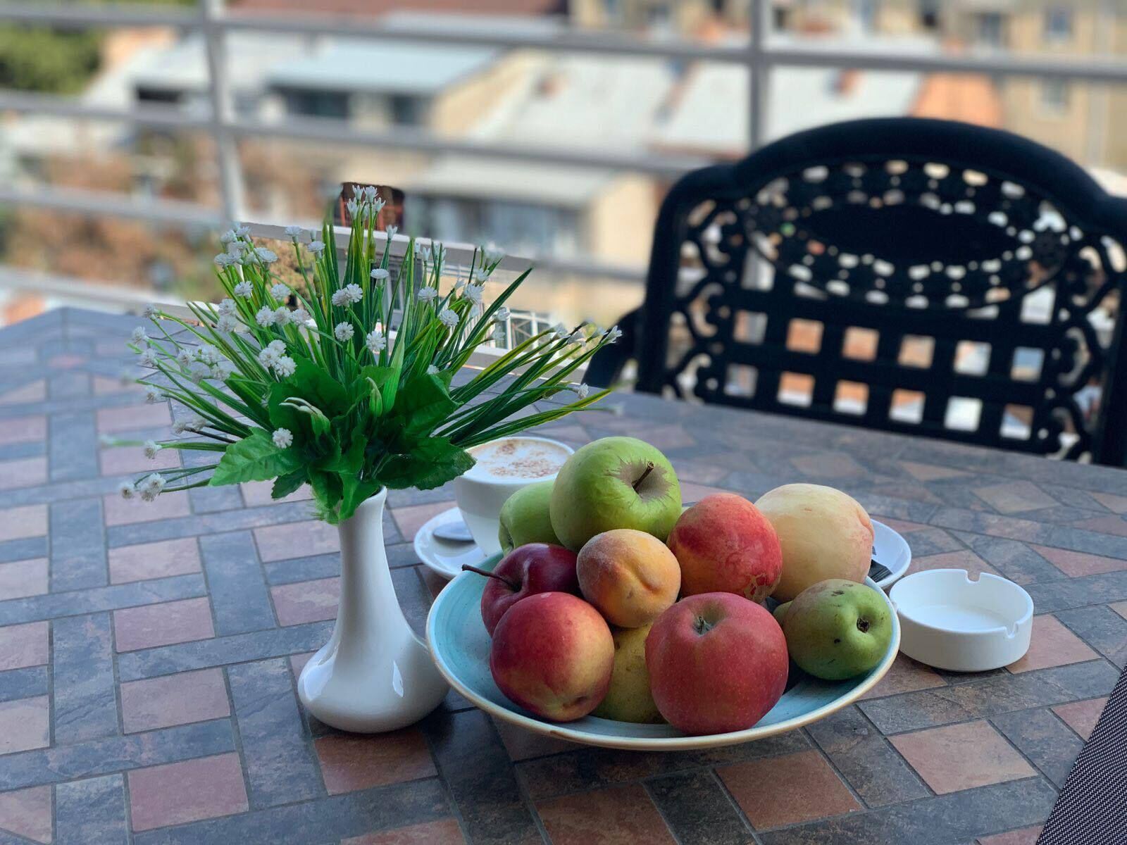 Una mesa con un plato de frutas, un florero con flores y un cenicero blanco.