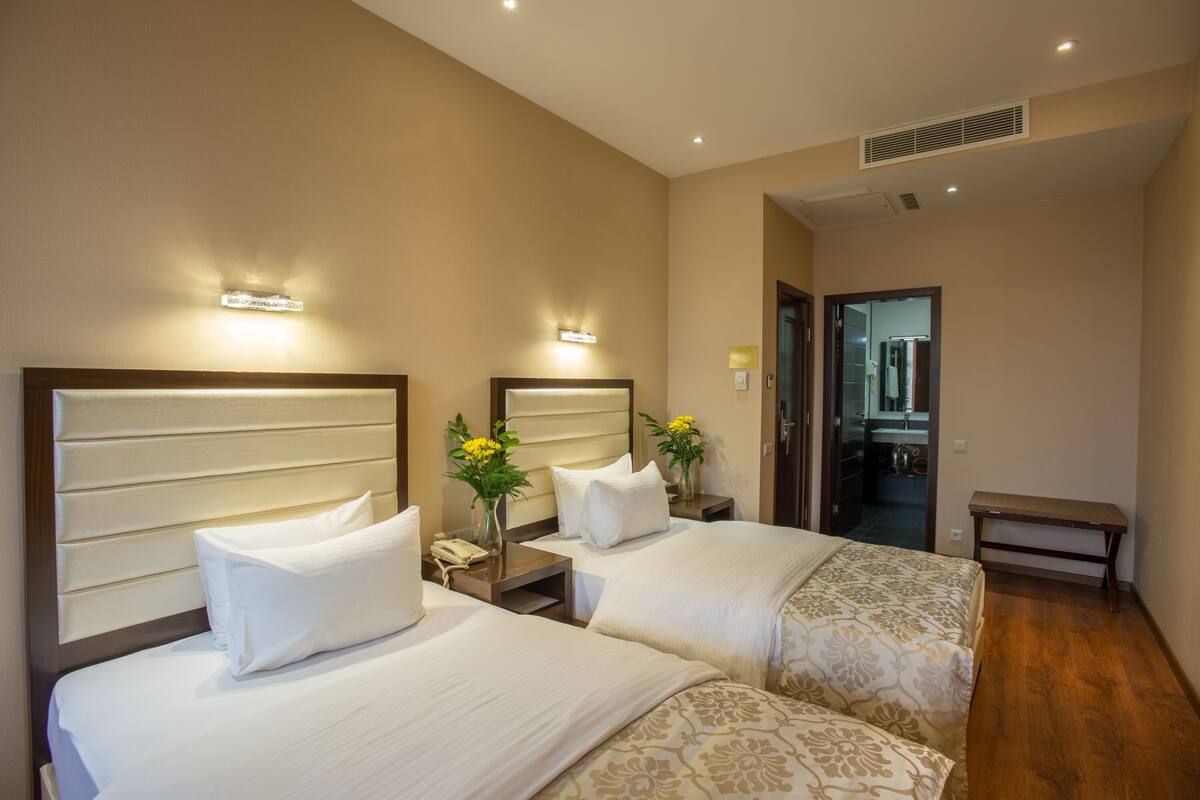 Habitación de hotel con dos camas, una mesita de noche con teléfono y un florero, y un banco.