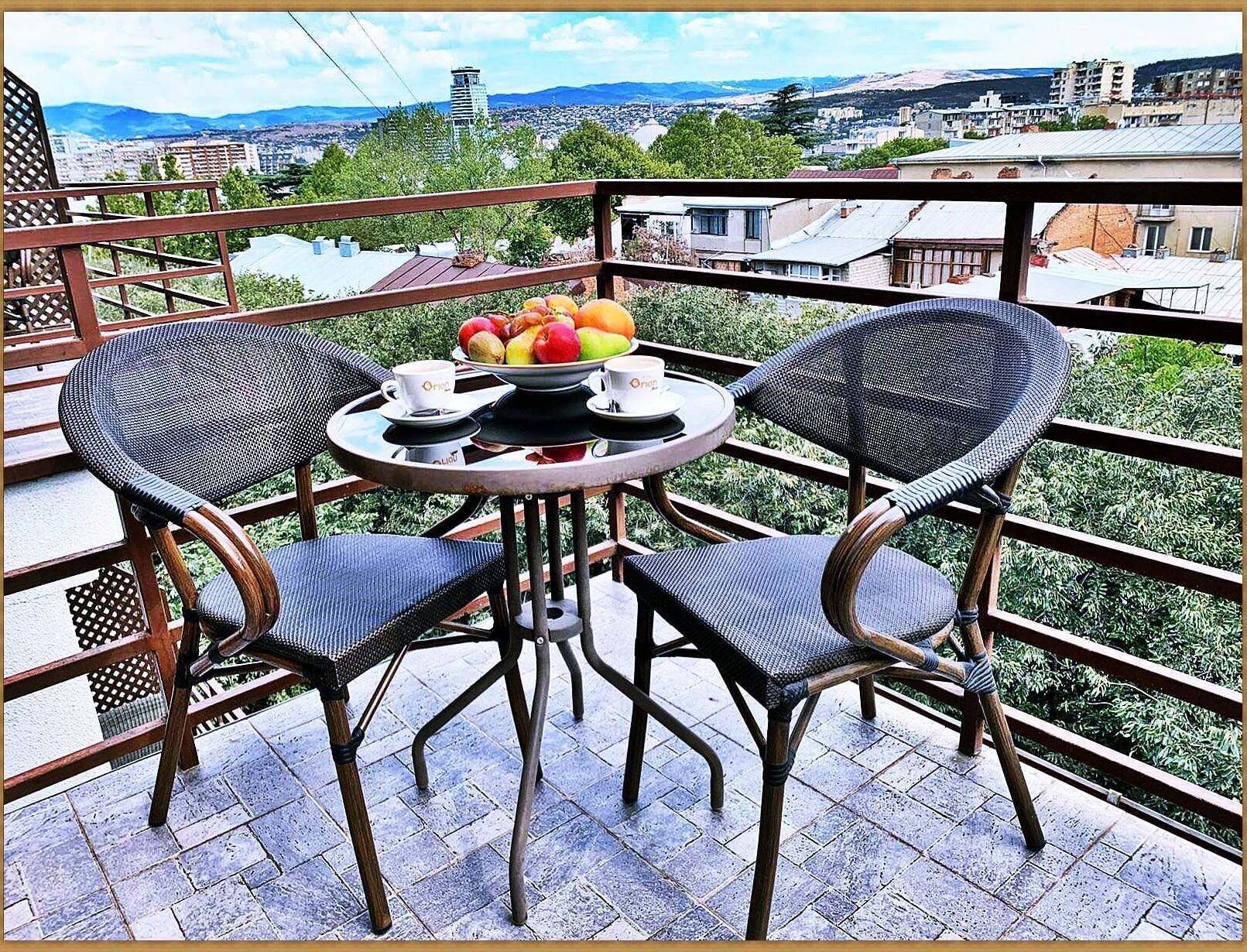 Ein Balkon mit zwei Korbstühlen, einem kleinen runden Tisch, einer Obstschale und zwei Kaffeetassen.