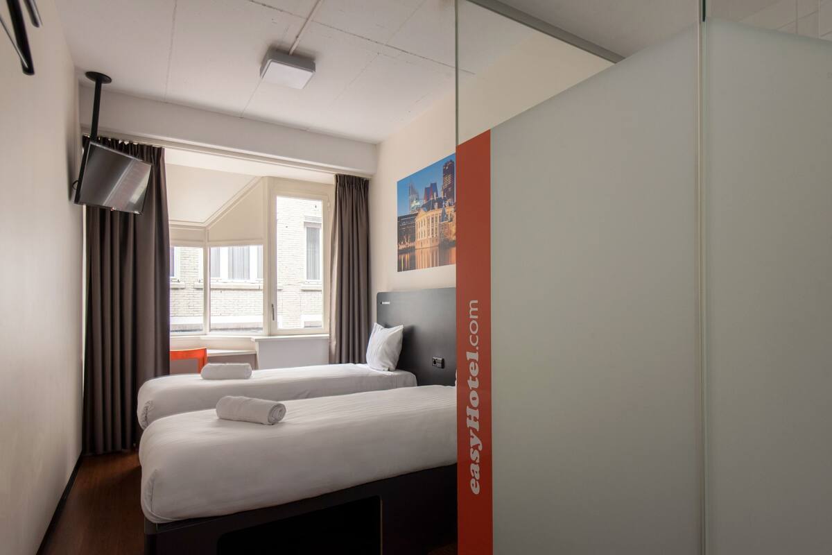 Habitación de hotel con dos camas, un televisor de pantalla plana, una ventana con cortinas y un cartel con un paisaje urbano en la pared.