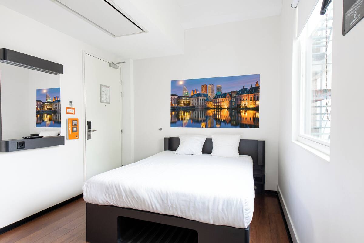 Una habitación de hotel moderna con una cama grande, un cuadro de paisaje urbano en la pared, una ventana con persianas y una puerta con un espejo y una mesita.