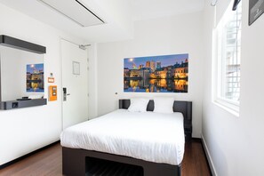 Une chambre d’hôtel moderne avec un grand lit, un tableau représentant un paysage urbain accroché au mur, une fenêtre avec des stores et une porte dotée d’un miroir et d’une petite table.