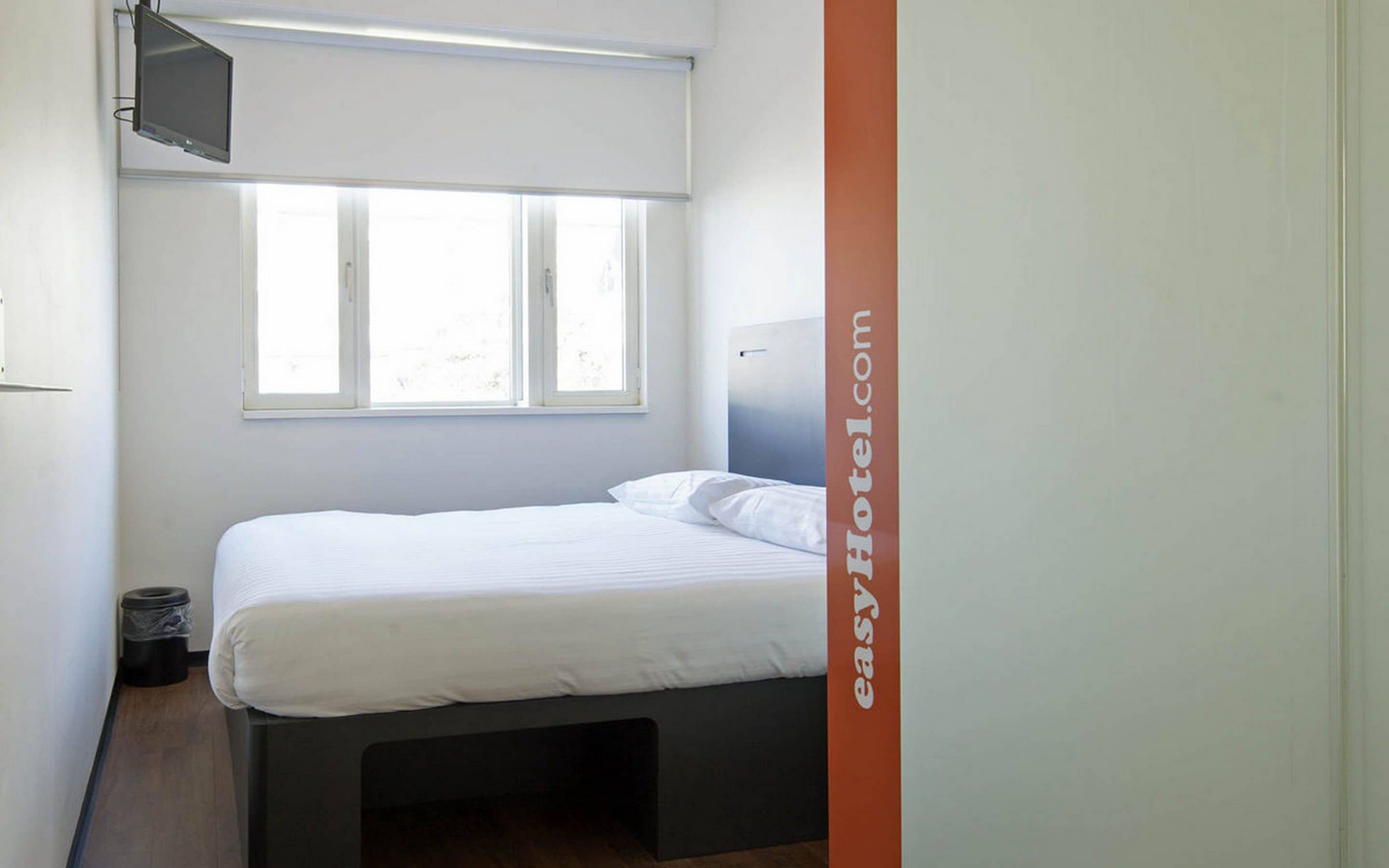 A single bed in a room with a window, a television on the wall, and a trash bin.