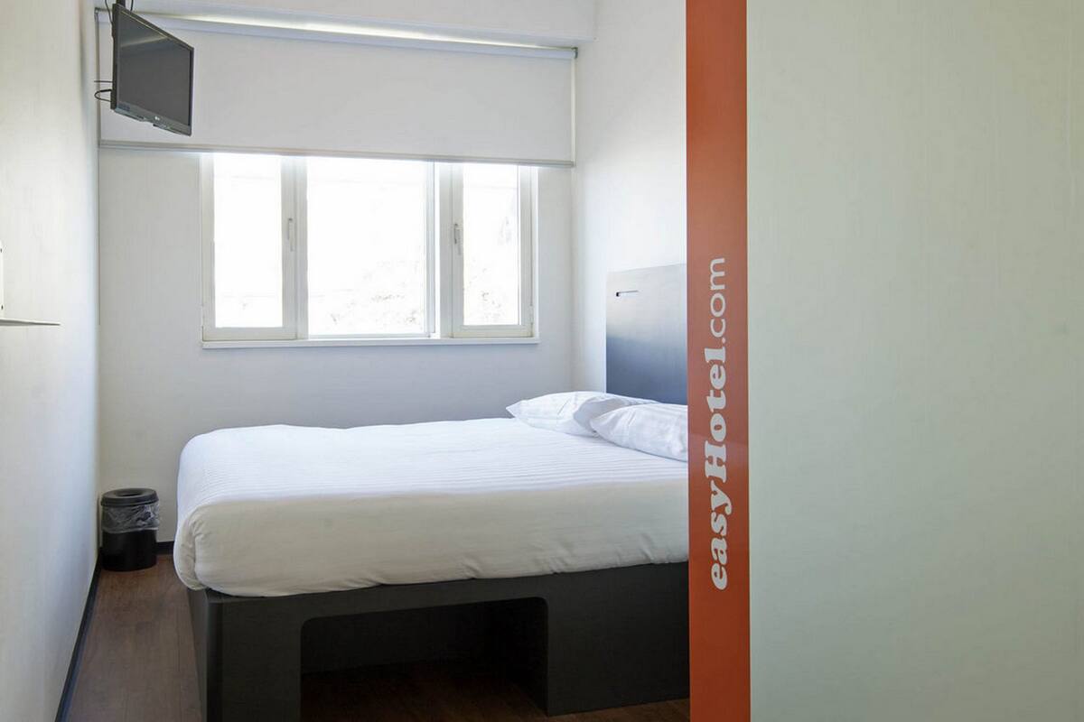 Una cama individual en una habitación con ventana, televisión en la pared y un cubo de basura.
