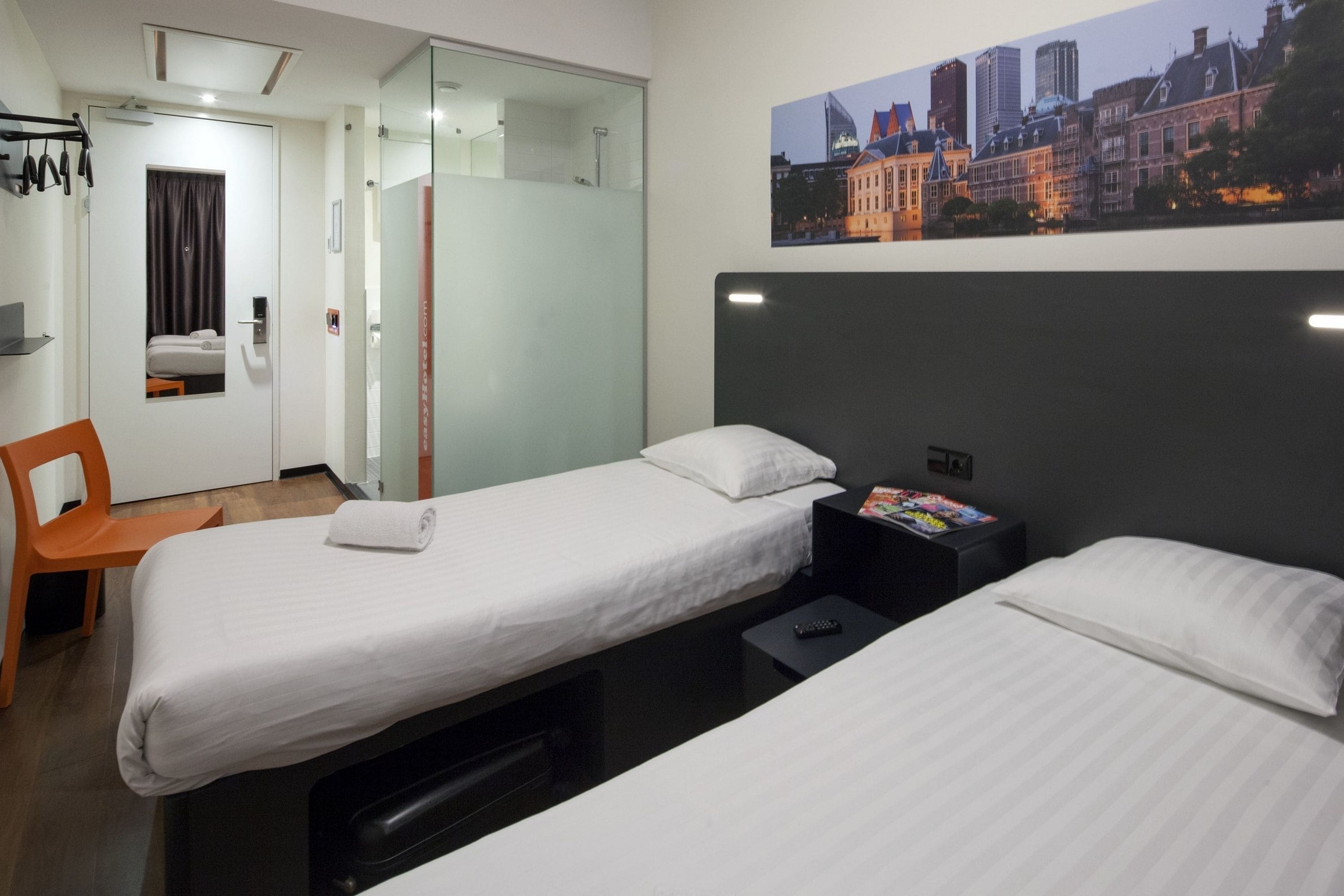 A hotel room with two beds, a desk, a chair, and a cityscape painting on the wall.