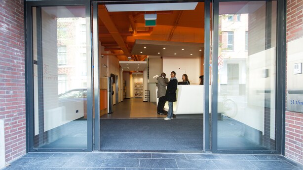 Una entrada de edificio moderna con puertas de cristal, un mostrador de recepción y una escalera visible que conduce al interior.