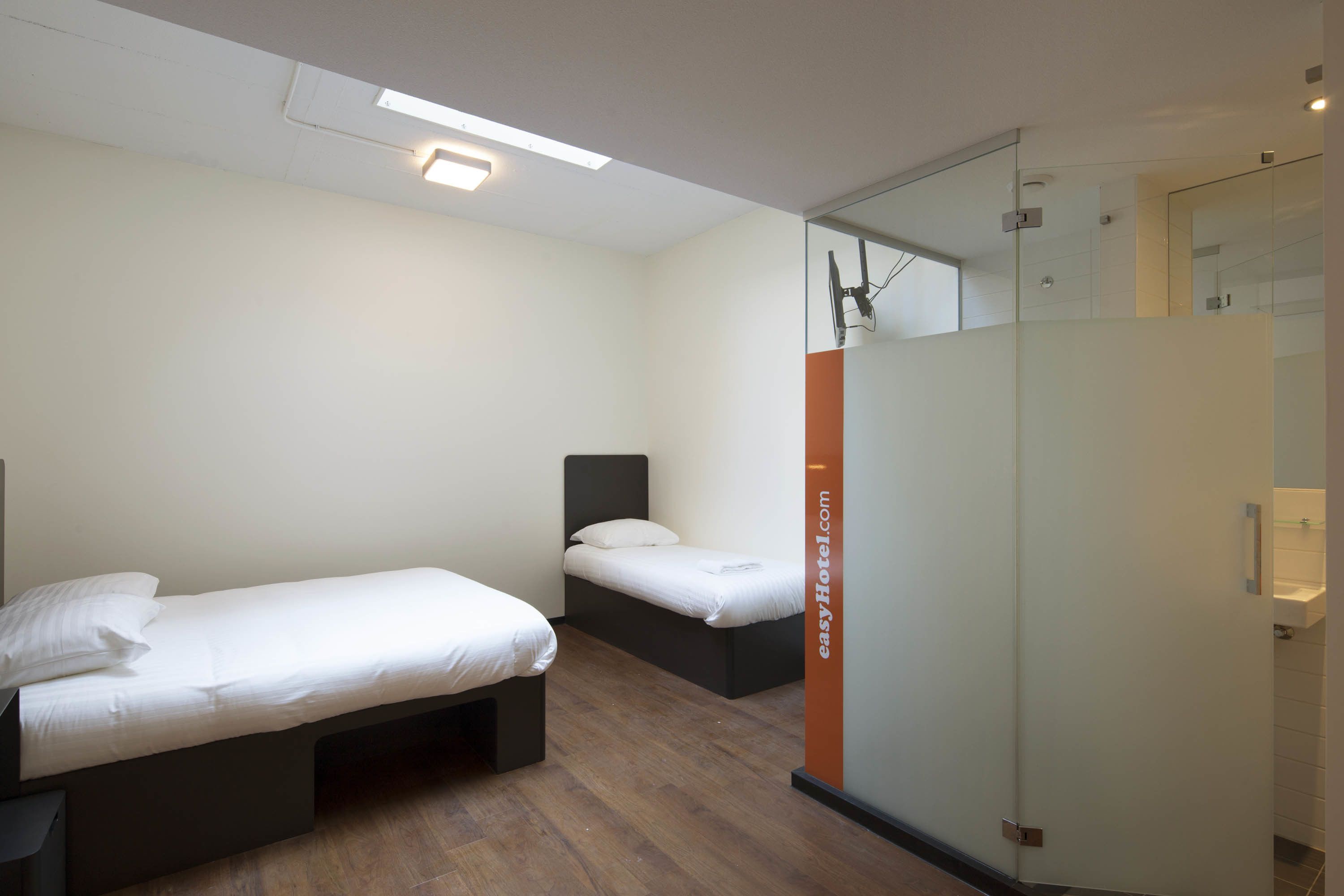 Une chambre d’hôtel moderne avec deux lits simples, une cloison en verre et une salle de bain visible.