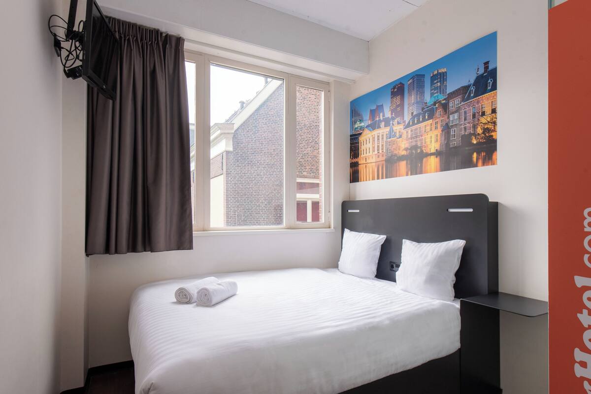 Una habitación de hotel con cama, ventana con cortinas, un cuadro de un paisaje urbano y una mesita de noche.