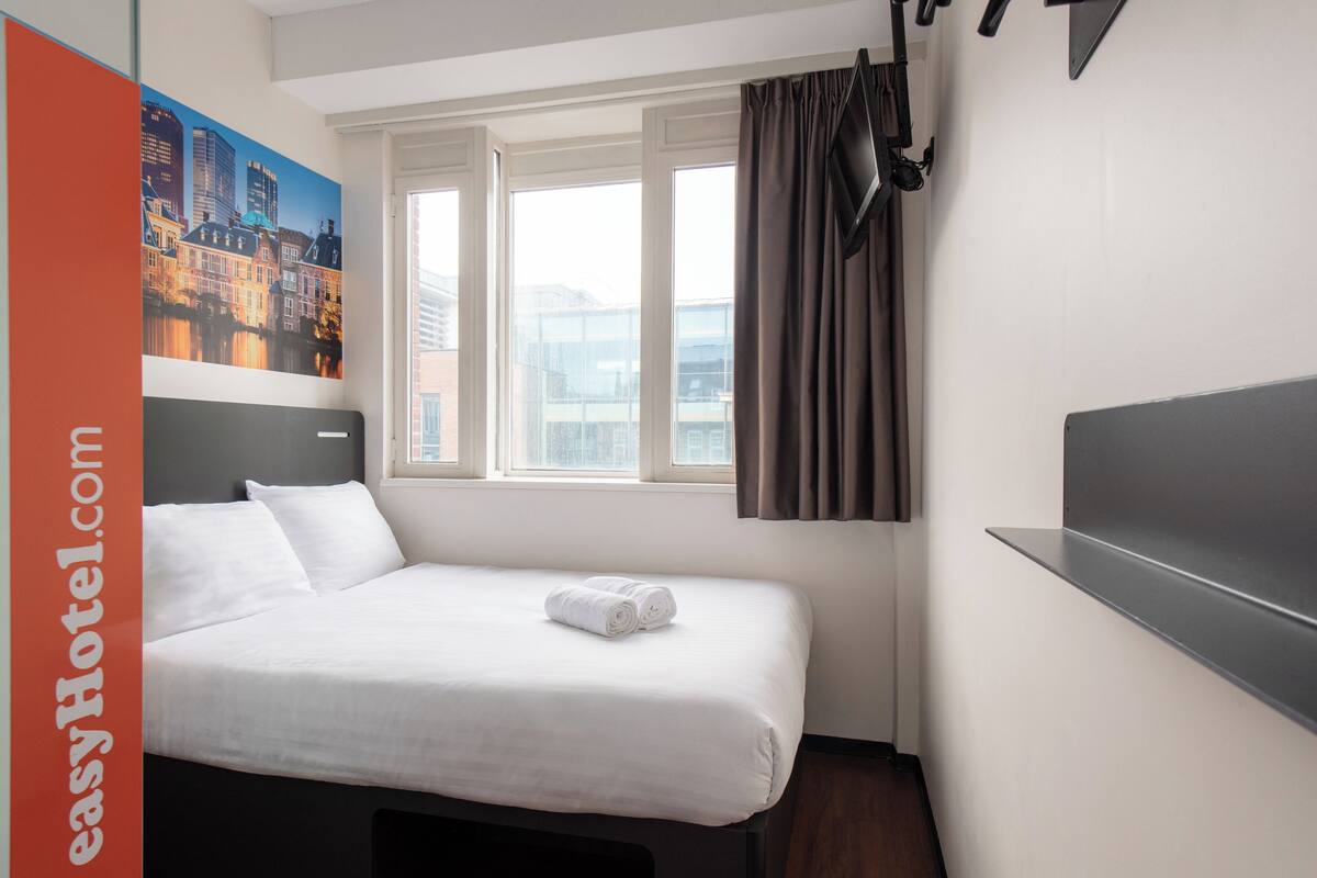 Habitación de hotel con cama, ventana con cortinas, televisor de pantalla plana y un cuadro de paisaje urbano en la pared.