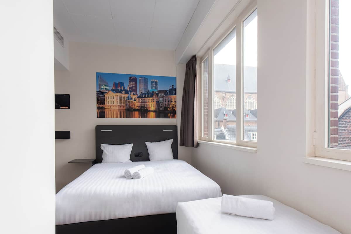 Habitación de hotel con una cama grande, una cama más pequeña, un cuadro de un paisaje urbano, una ventana con cortinas y un televisor de pantalla plana en la pared.