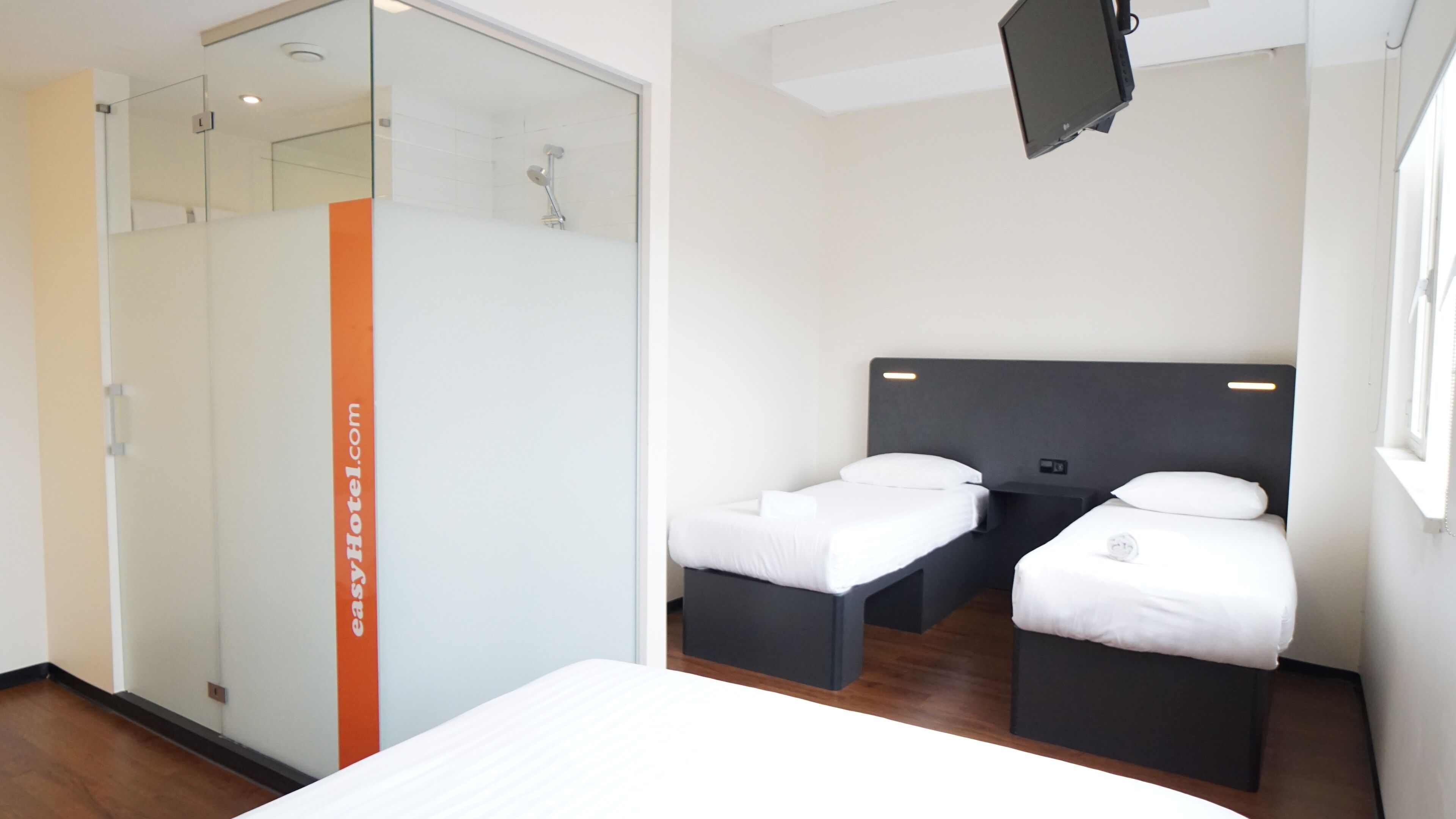 A modern hotel room with a large bed, two smaller beds, a glass shower enclosure, and a television mounted on the wall.