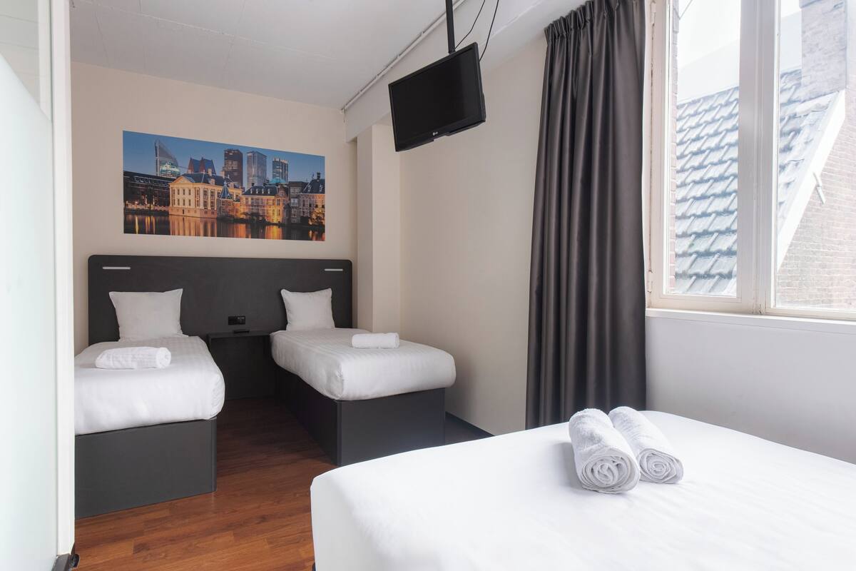Habitación de hotel con dos camas, un televisor de pantalla plana, un cuadro de un paisaje urbano y una ventana con cortinas.