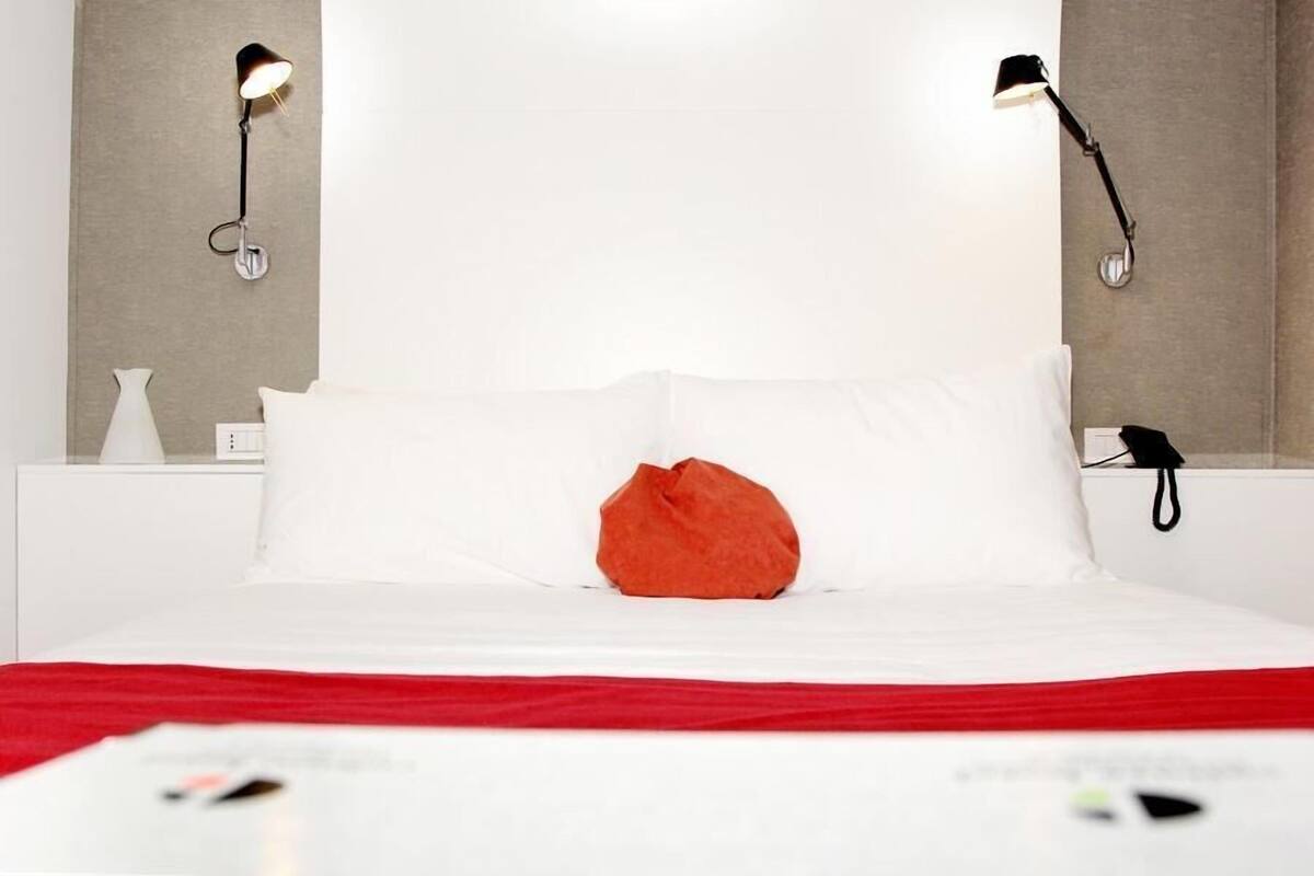 A bed with white bedding, a red throw pillow, and two wall-mounted lamps.
