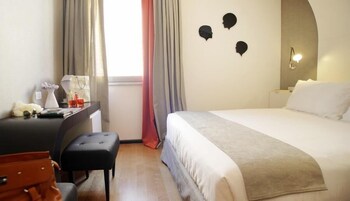 Hotelkamer met een bed, een bureau met stoel, een kleine tafel met lamp, en een muur met drie zwarte silhouetten.