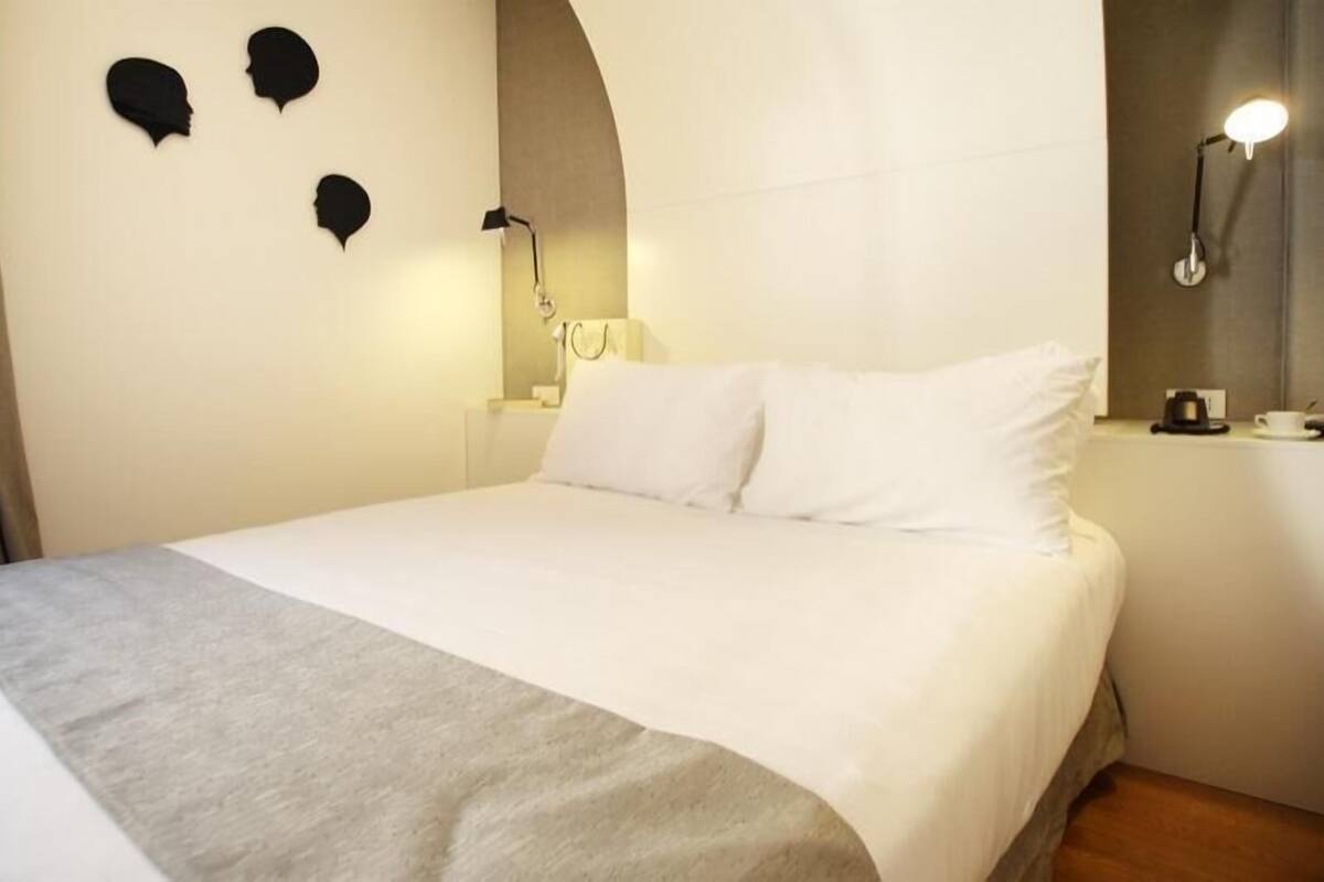 A hotel room with a bed, a nightstand, a lamp, and a wall-mounted light fixture.