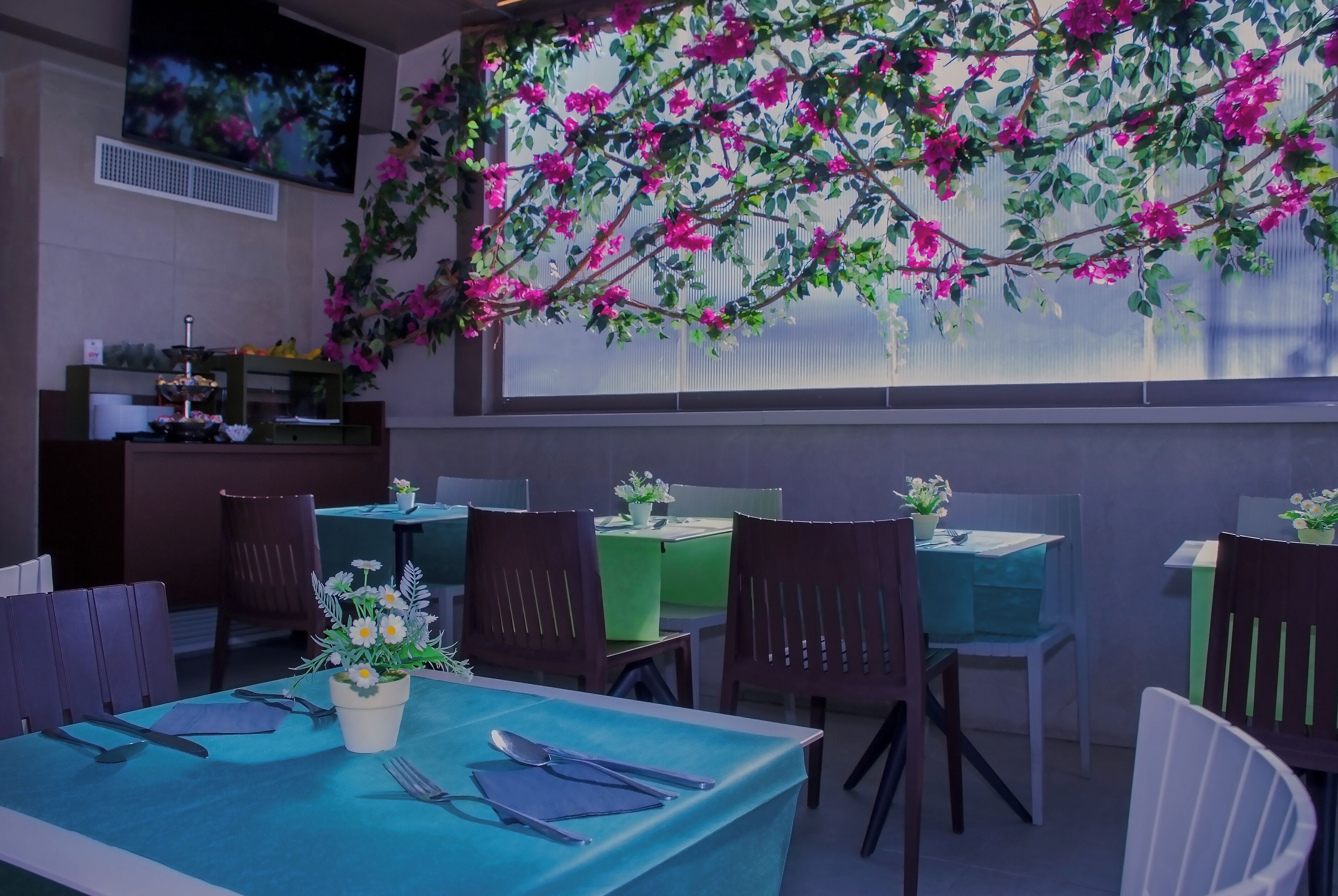 A restaurant with tables set for dining, chairs, a TV screen, and a decorative plant with pink flowers.