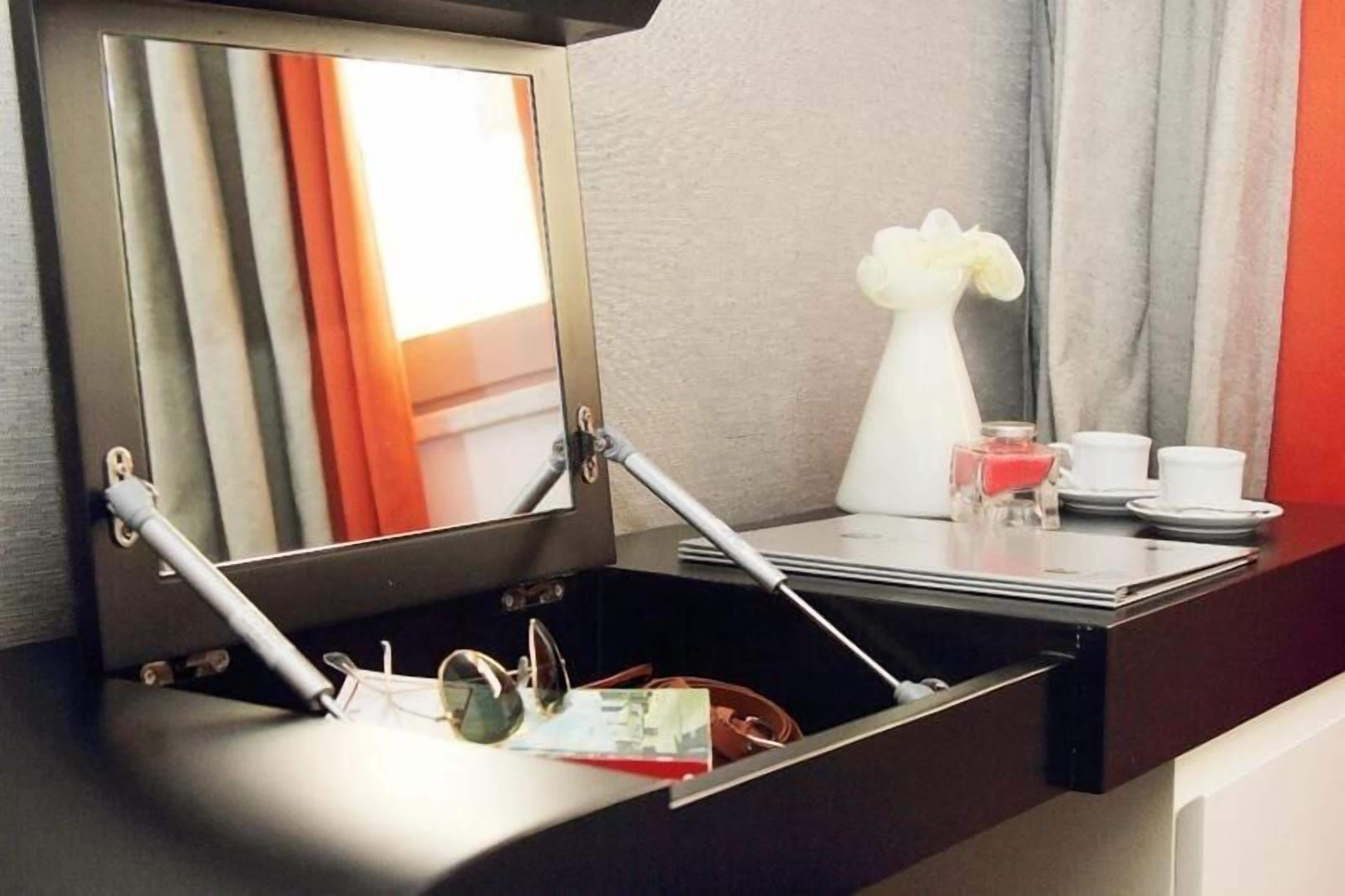 A hotel room with a mirror, a small table with a vase, and a drawer with sunglasses and a wallet.