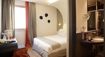 Una camera d'albergo con un letto, una scrivania con una sedia, una valigia e un bagno con uno specchio e un lavandino.