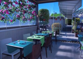 An outdoor dining area with tables and chairs, covered by a roof, and surrounded by greenery and flowers.