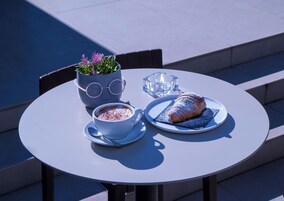Una mesa redonda con una taza de chocolate caliente, un croissant y una planta en maceta con gafas encima.