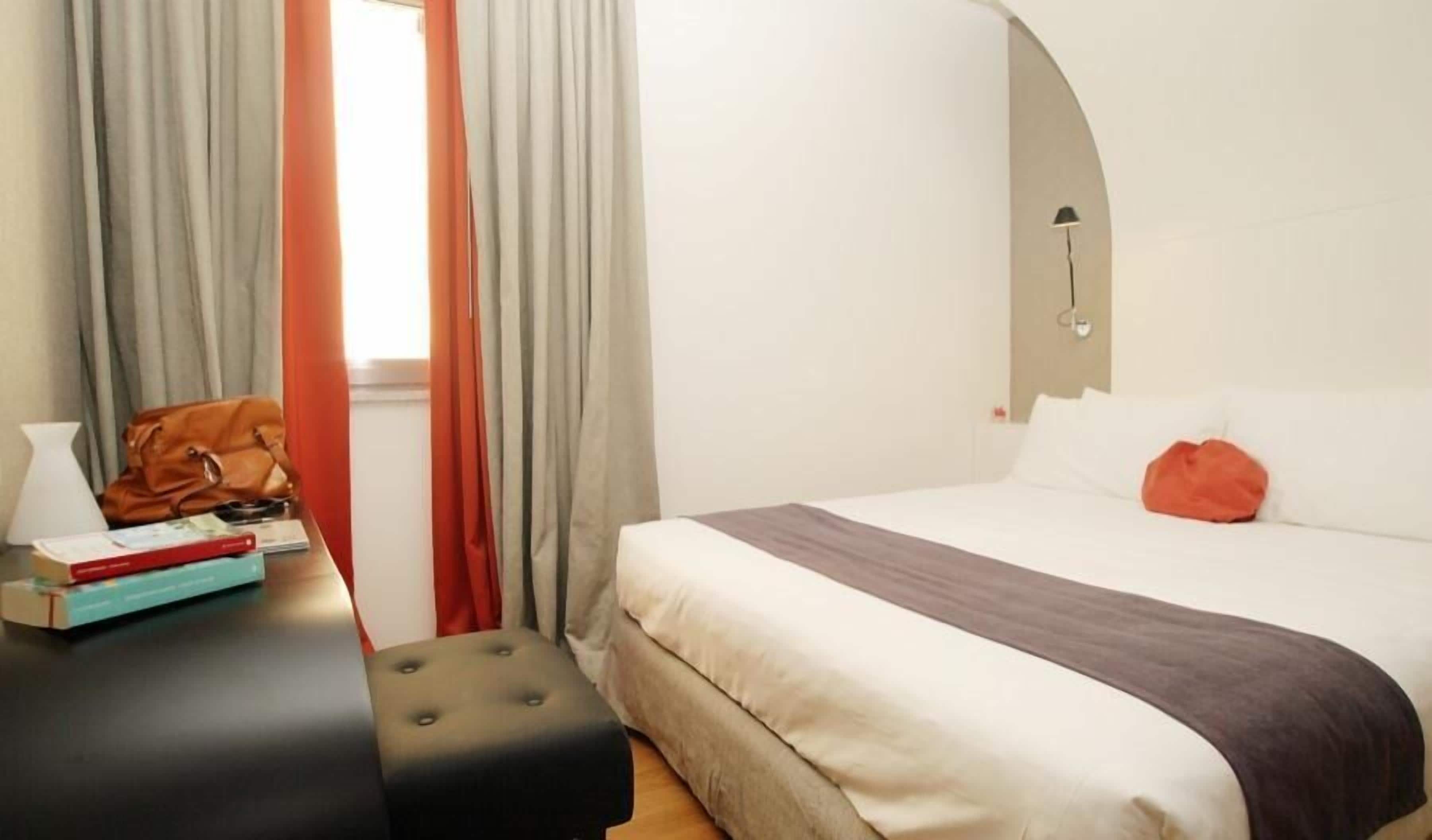 A hotel room with a bed, a nightstand, a lamp, and a window with curtains.