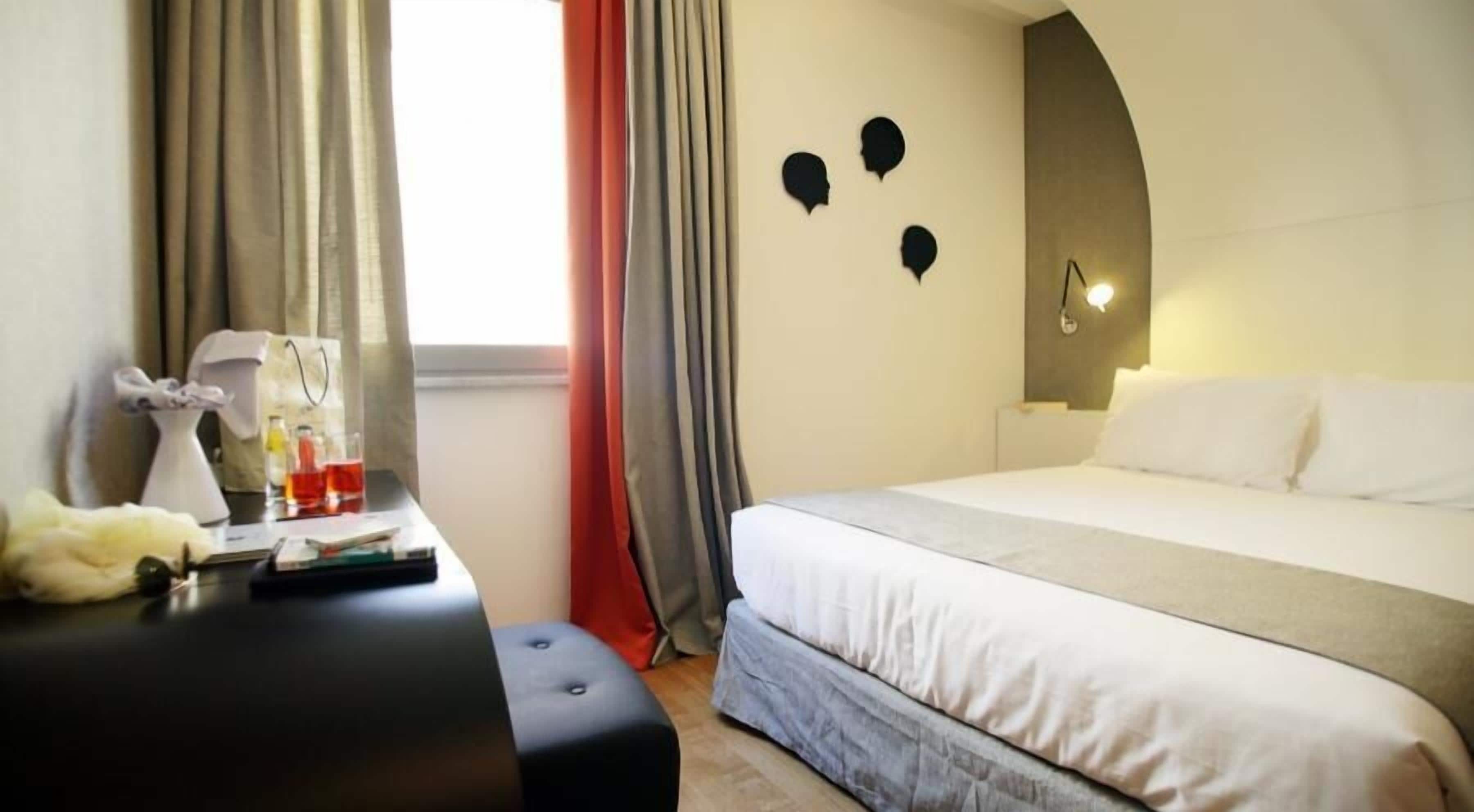 A hotel room with a bed, a desk with a chair, a vase, and a window with curtains.