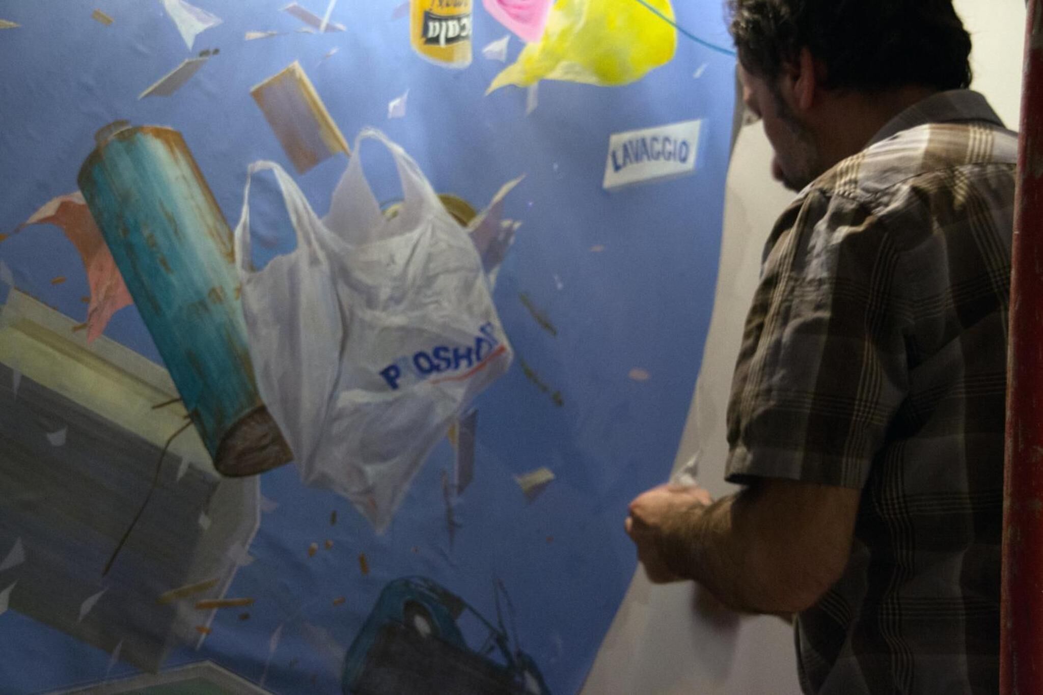 A man standing in front of a wall with a large poster featuring a blue background, a white plastic bag, and some text.