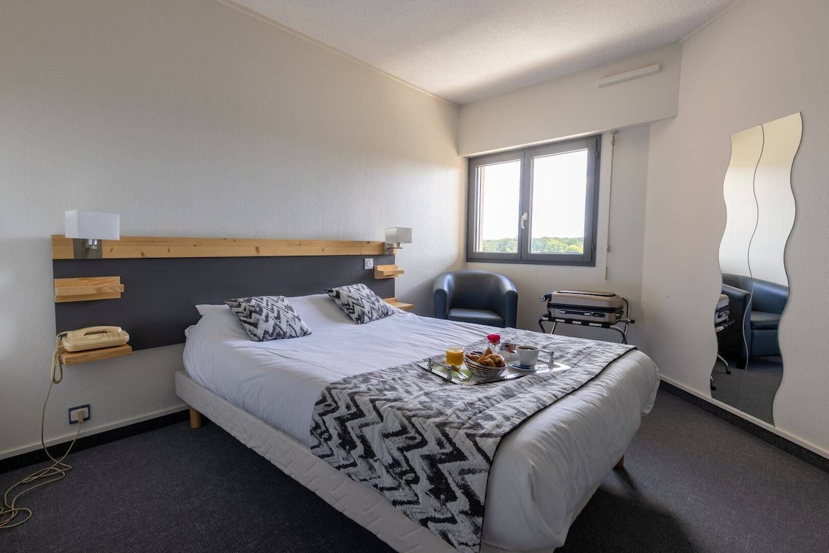 A hotel room with a bed, bedside table, lamp, chair, and a window.
