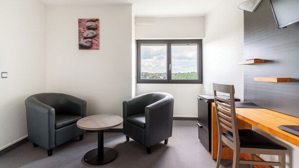 A modern room with two armchairs, a small round table, a wooden desk, and a TV mounted on the wall. - Abor Hôtel (Saint-Pierre-du-Mont)