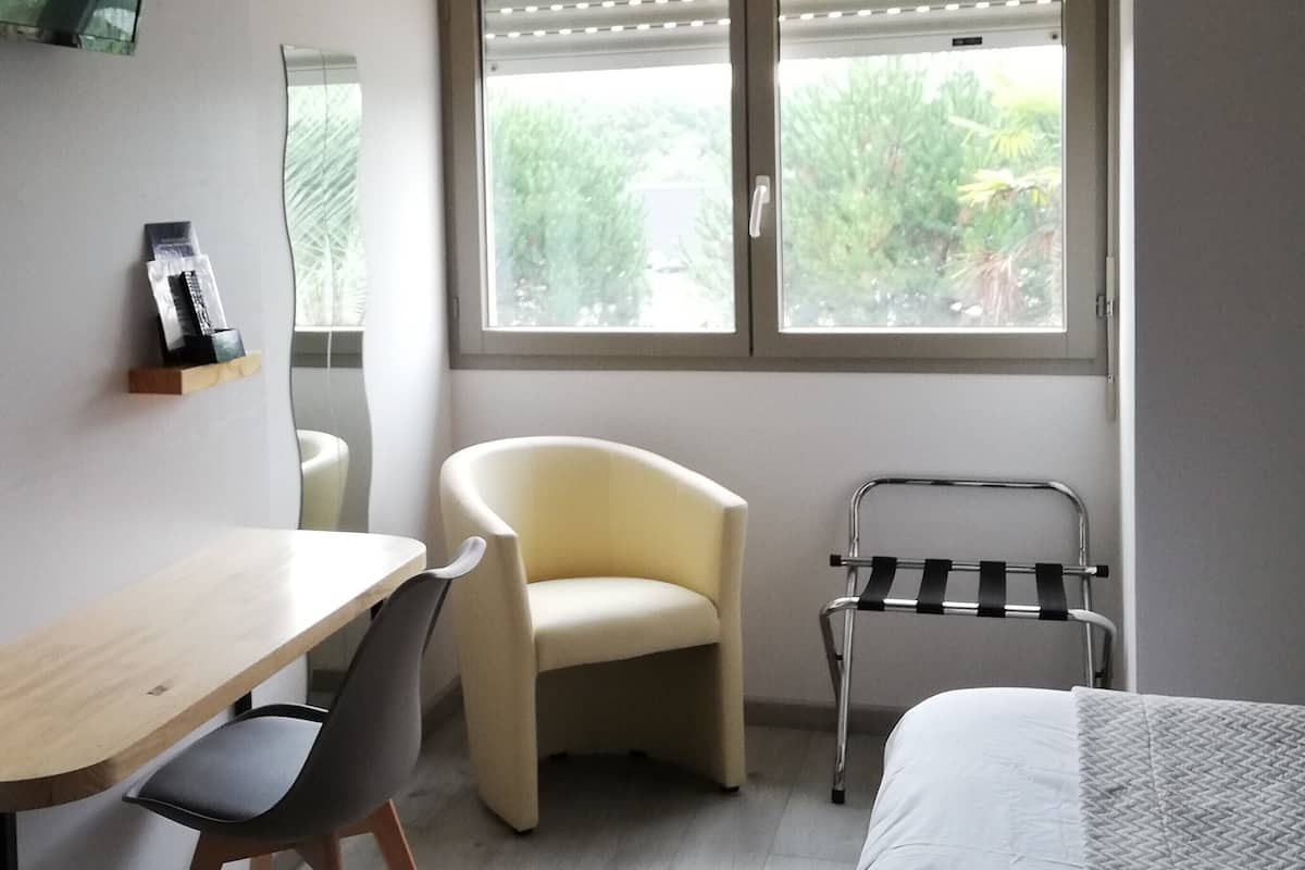 A bedroom with a bed, a desk, a chair, a small table, a trash can, and a window with blinds.