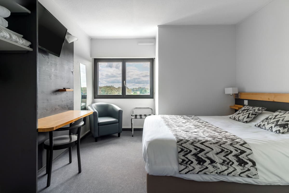 A modern hotel room with a bed, a desk, a chair, and a window with a view of greenery.
