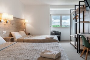 A hotel room with two beds, a desk, a chair, a TV, and a window with a view of greenery.