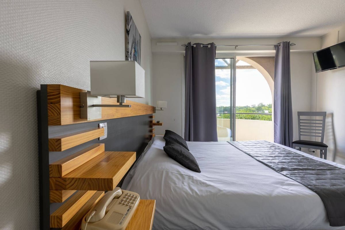 A hotel room with a bed, a desk with a telephone, a TV mounted on the wall, and a balcony with curtains.